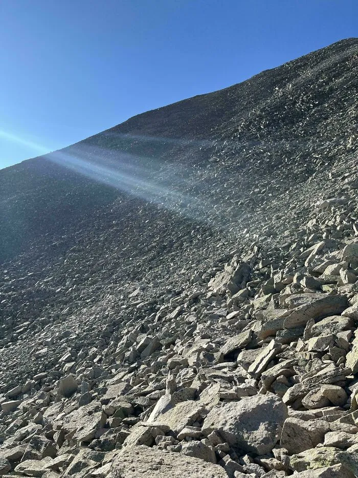 Rocky hillside under blue sky with sunlight beams, featuring perfectly camouflaged lizard blending with the stones.