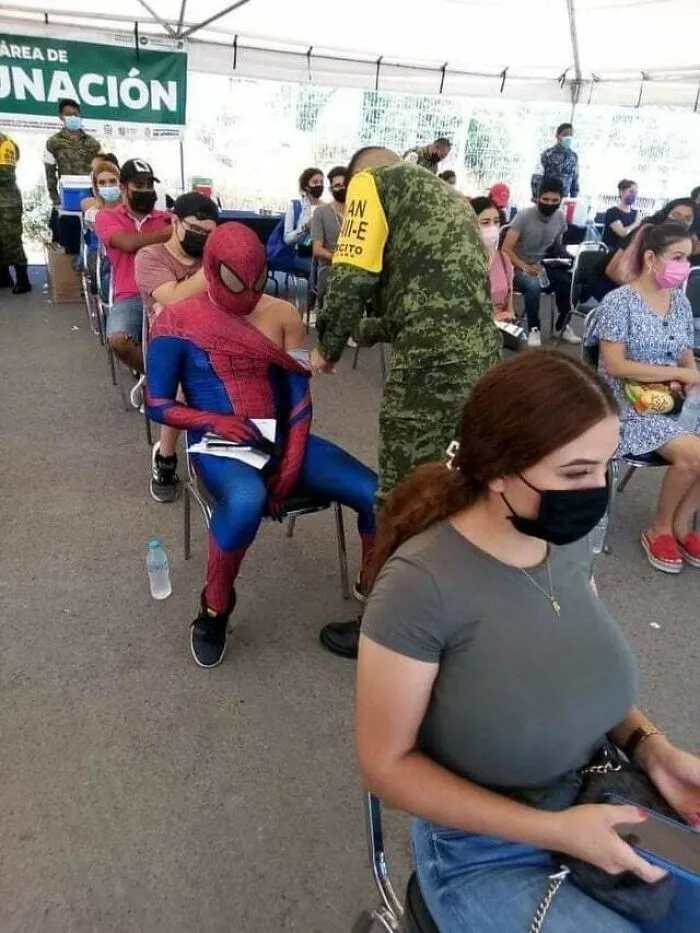 Person dressed as Spider-Man receiving a vaccine shot in a crowded vaccination area with others waiting in line.