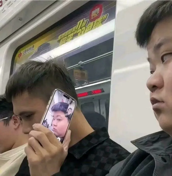 Man holding phone showing someone else's face to his ear on subway, a weird and confusing cursed image.