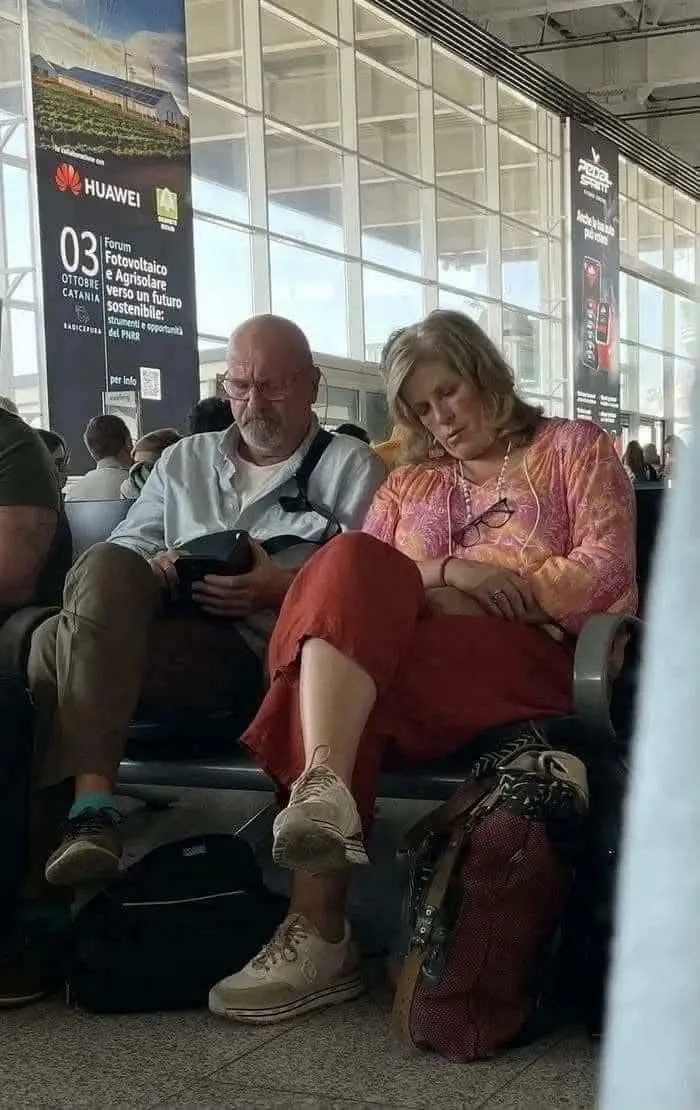 Two travelers at an airport waiting area, one sleeping and the other using a device, creating a weird and confusing scene.