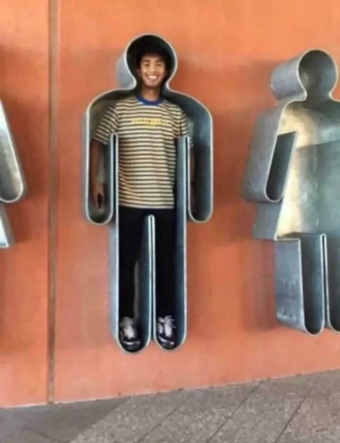 Person standing inside a metal figure sculpture, creating a weird and confusing optical illusion outdoors on a wall.