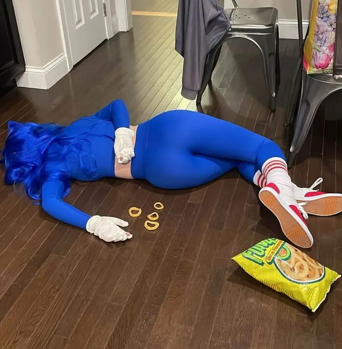 Person in blue suit and wig lying face down on floor with snack chips scattered nearby in a weird confounding scene.