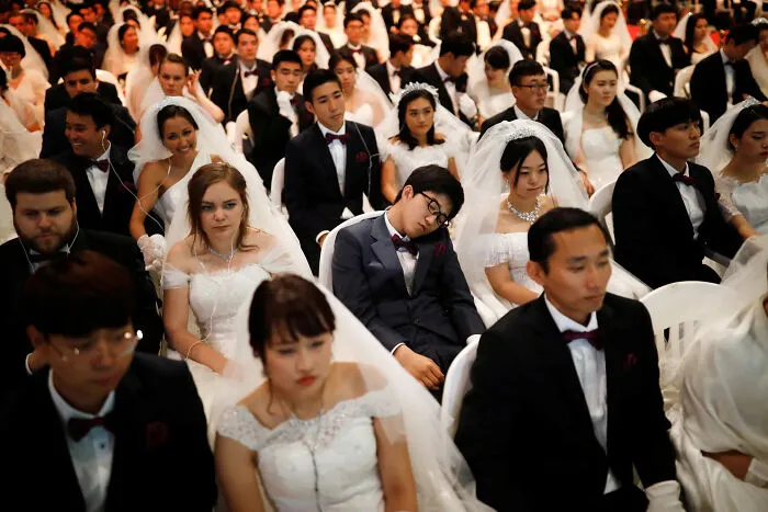 Hundreds of brides and grooms in formal wedding attire seated closely in a large strange and confusing group photo.