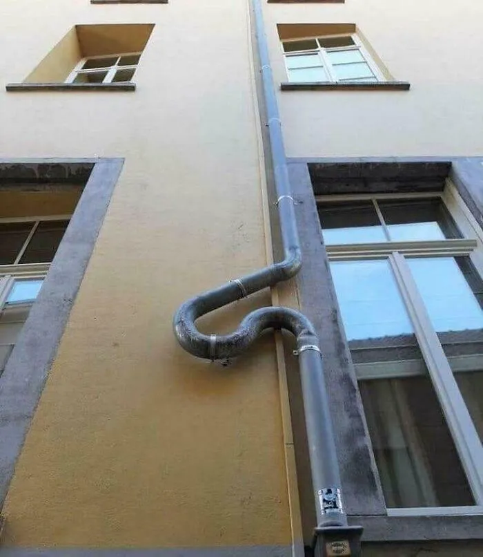 Curved metal pipe on building wall creating a weird and confusing visual in an urban outdoor setting.