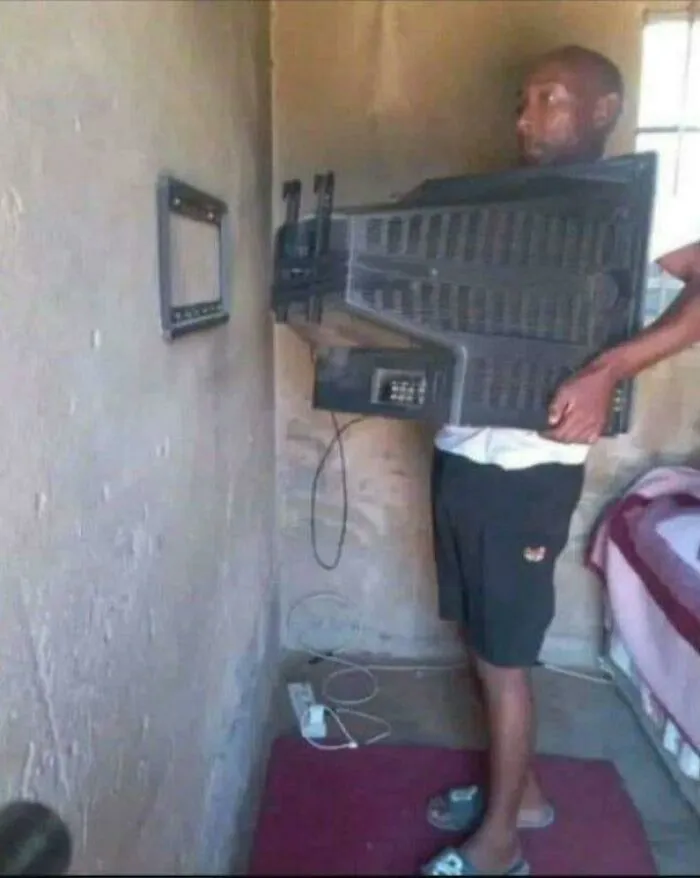 Man holding a large TV to his chest while facing a tiny wall-mounted screen in a weird and confusing cursed image.