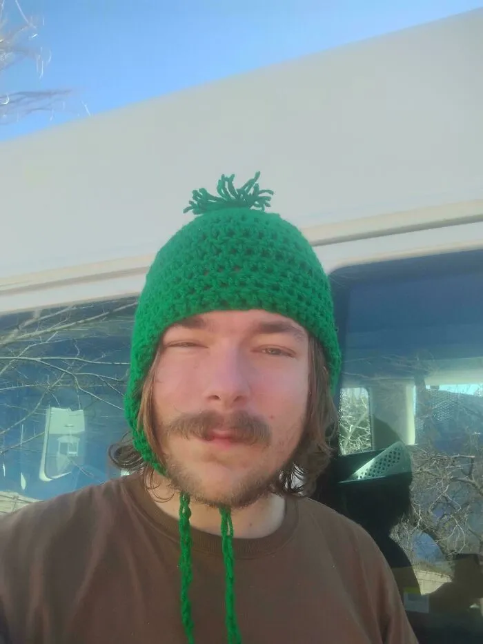 A person wearing a green knitted beanie with earflaps and a pom-pom, a cool find from dumpster diving.
