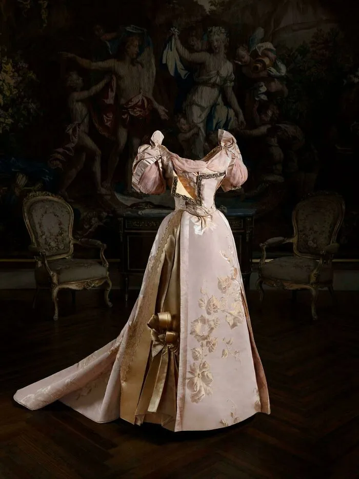 Elegant historical garment in pale pink with floral embroidery displayed in an ornate room, showcasing vintage fashion details.