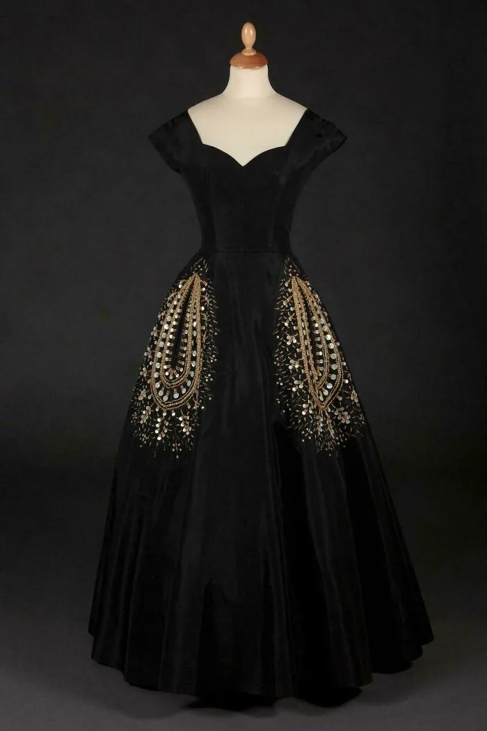 Black historical garment with gold and silver sequin embroidery displayed on a mannequin against a dark background.