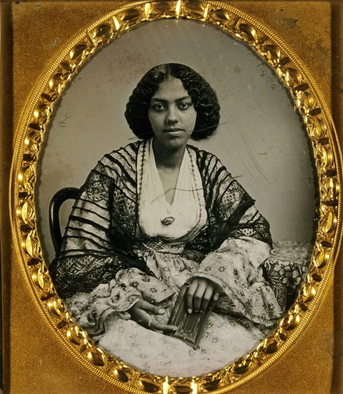 Victorian-era woman wearing intricate historical garments with lace and floral patterns in an ornate gold frame portrait.
