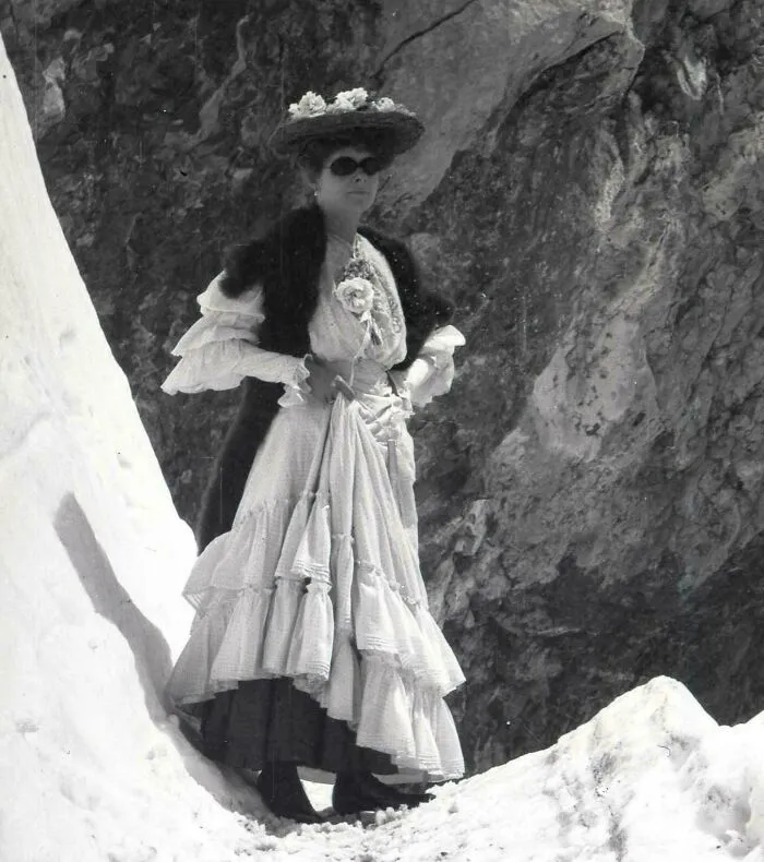 Woman wearing historical garments with layered dress and fur shawl, standing on snowy rocky terrain in vintage style.