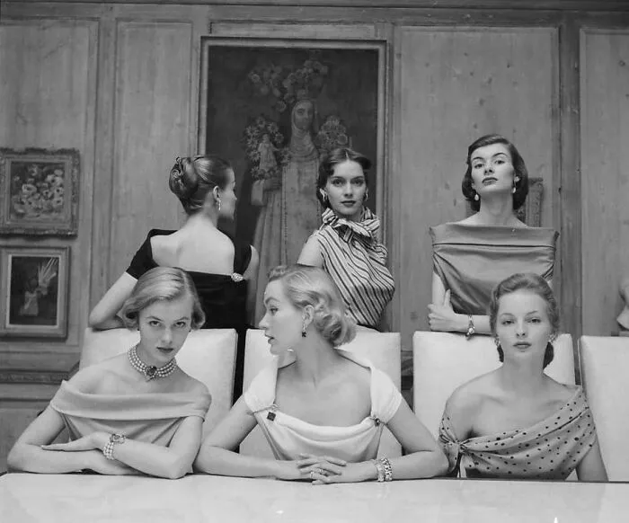 Six women in elegant vintage gowns and jewelry pose in a classic setting showcasing historical garments fashion.