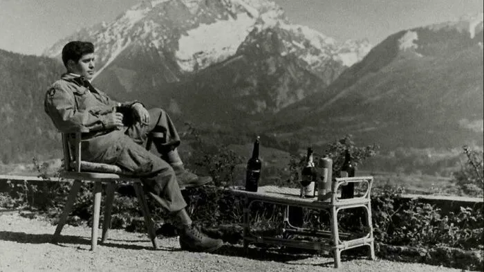 Man in military uniform sitting outdoors with bottles on a table, captured in a historical photo from a vastly different world.