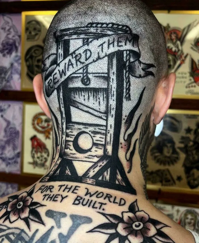 A stunning tattoo of a guillotine with text on the back of a person's head, surrounded by other stunning tattoos.