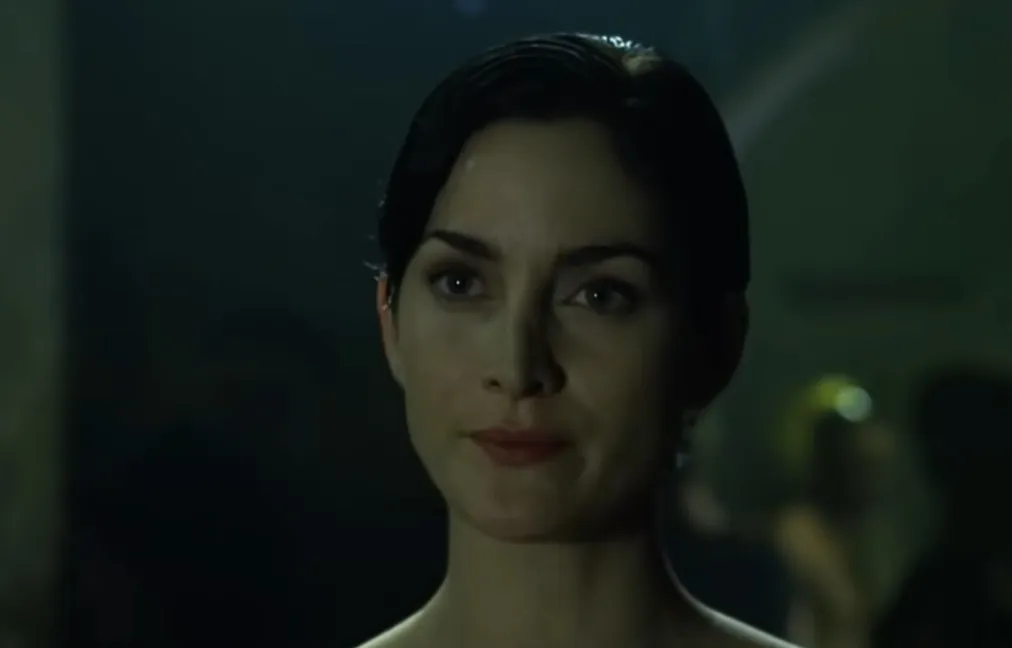 Close-up of Trinity from The Matrix with short dark hair, looking thoughtfully in a dimly lit environment.