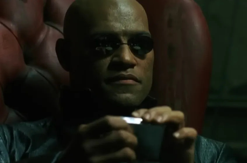 Morpheus from The Matrix wearing black sunglasses, seated in a red chair, holding a small black object.