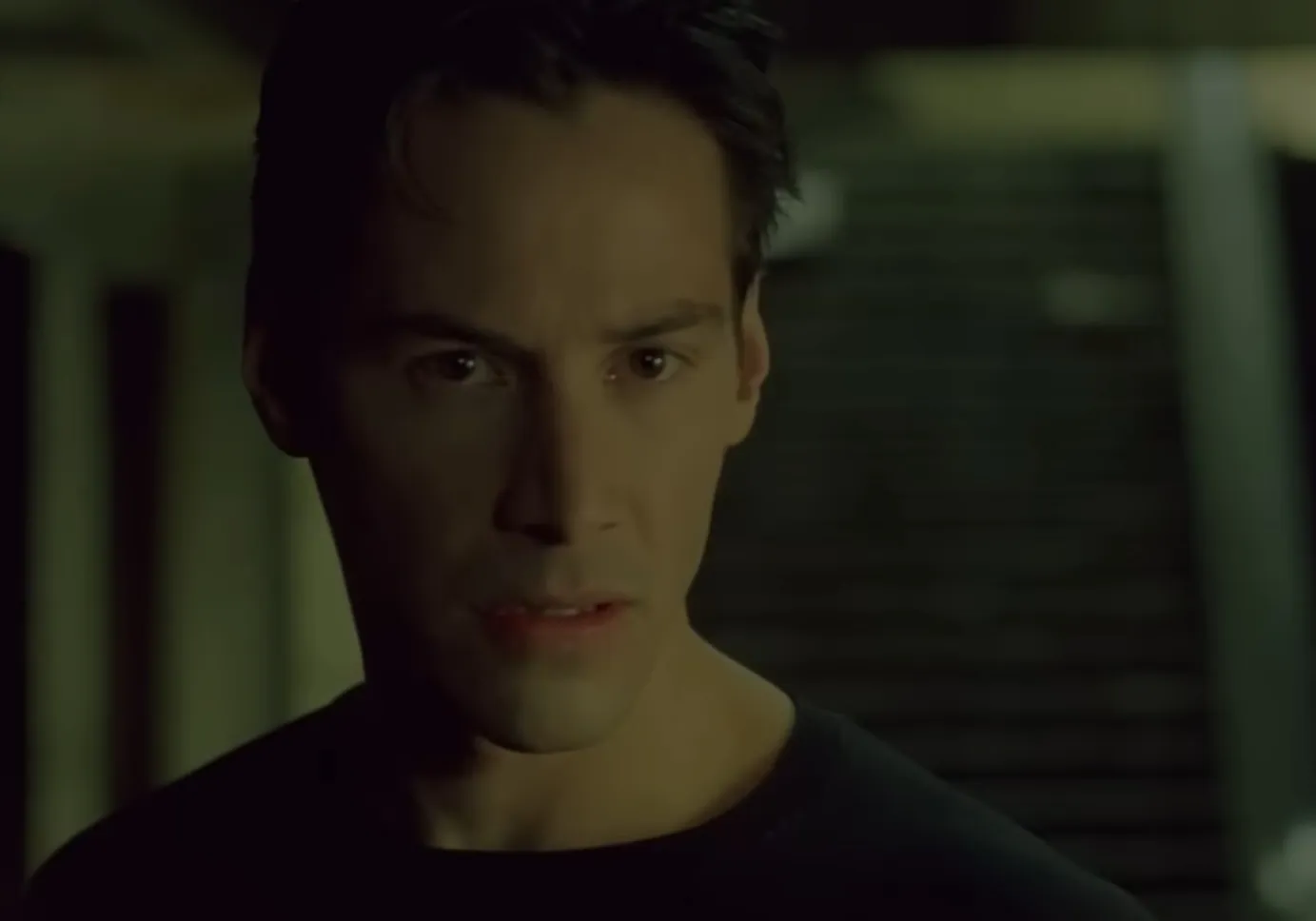 Close-up of Neo from The Matrix with a serious expression in a dimly lit background scene.