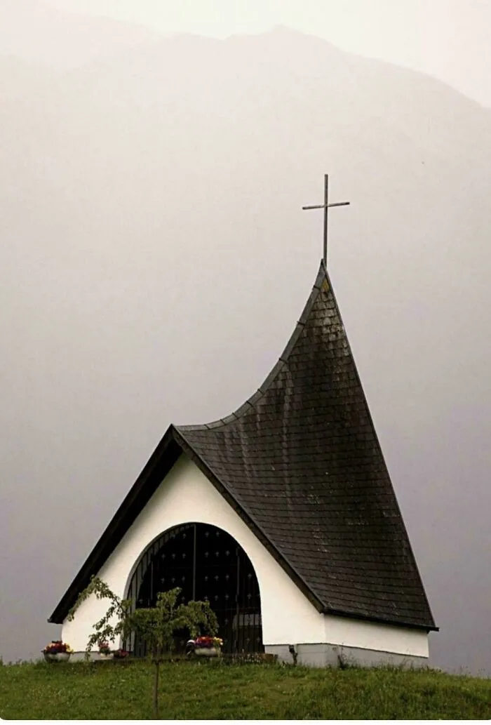Small, beautiful architecture of a chapel with a dark, pointed roof and cross, set against a misty mountain background.