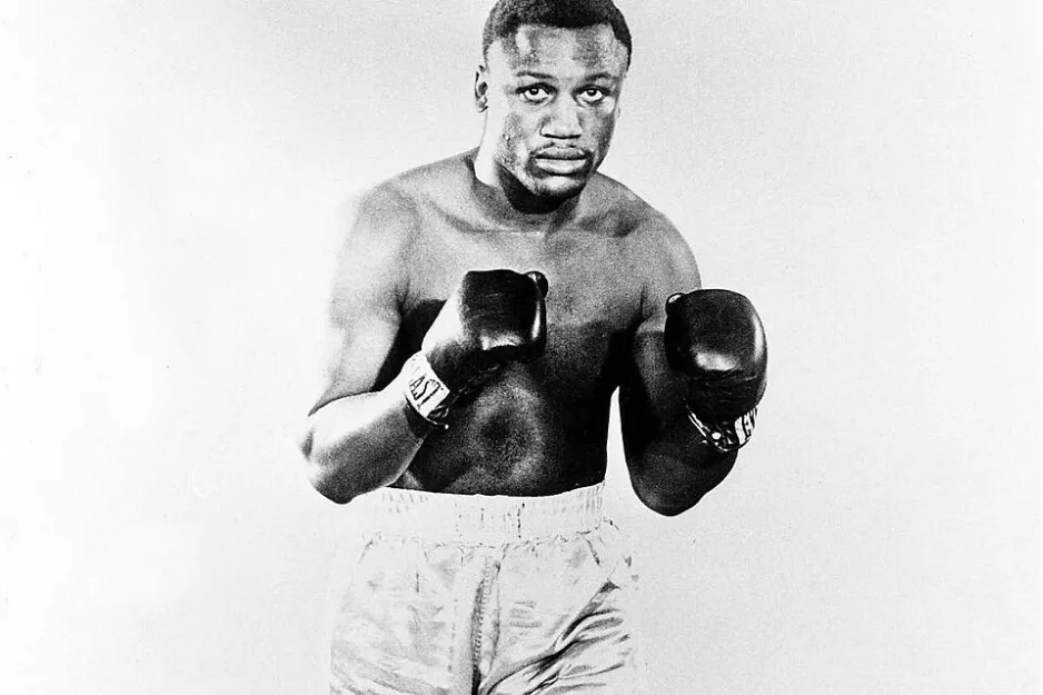 Joe Frazier in a boxing stance, wearing gloves and trunks, preparing for a famous 1974 boxing match.