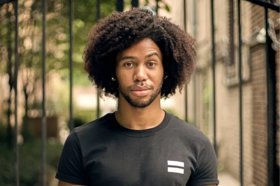 Young man with a large afro hairstyle standing outdoors, wearing a black t-shirt in a casual setting.