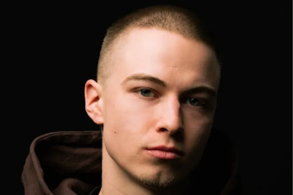 Young man with a buzz cut hairstyle, wearing a dark hoodie, photographed against a black background.