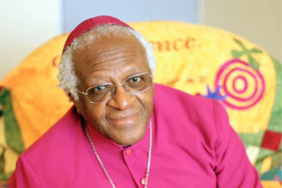 Desmond Tutu wearing glasses and a magenta religious robe, smiling gently in a colorful indoor setting.