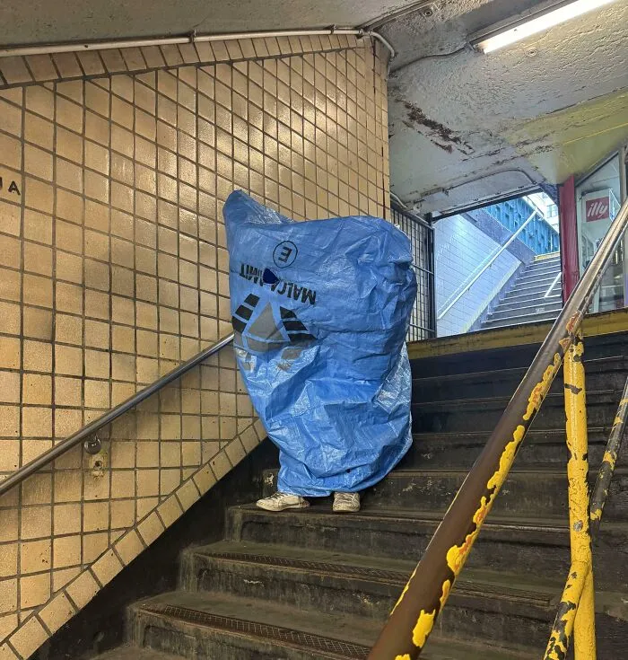 Person covered in a blue tarp standing on worn stairs inside a NYC subway station, showing a weird subway moment.