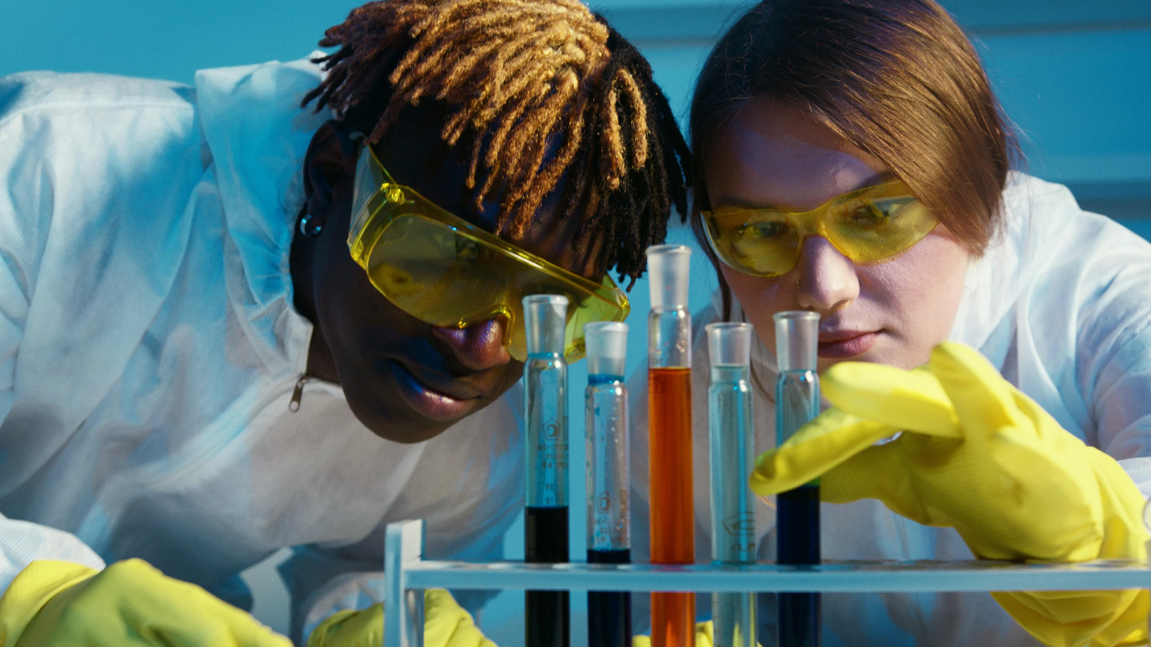 Two students in lab coats and yellow gloves perform a GCSE Combined Science quiz experiment with test tubes.
