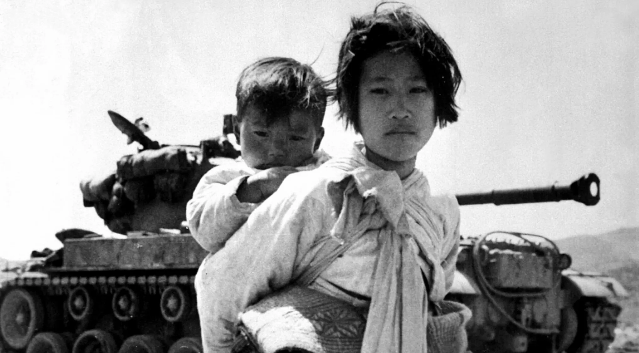 Young Korean girl carrying a child on her back near a military tank during the Korean War start period.