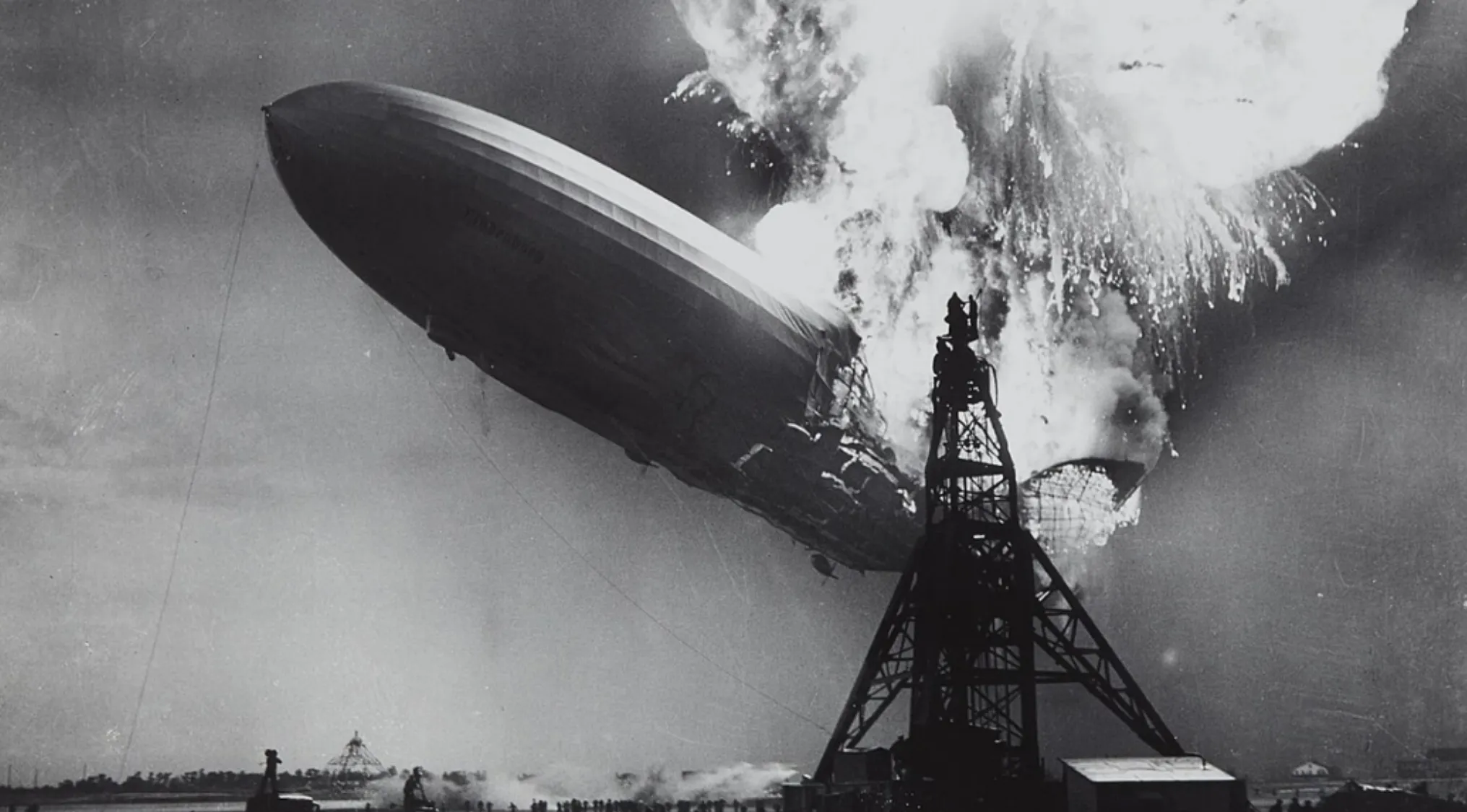 Hindenburg disaster airship engulfed in flames above mooring mast during 1930s disaster.