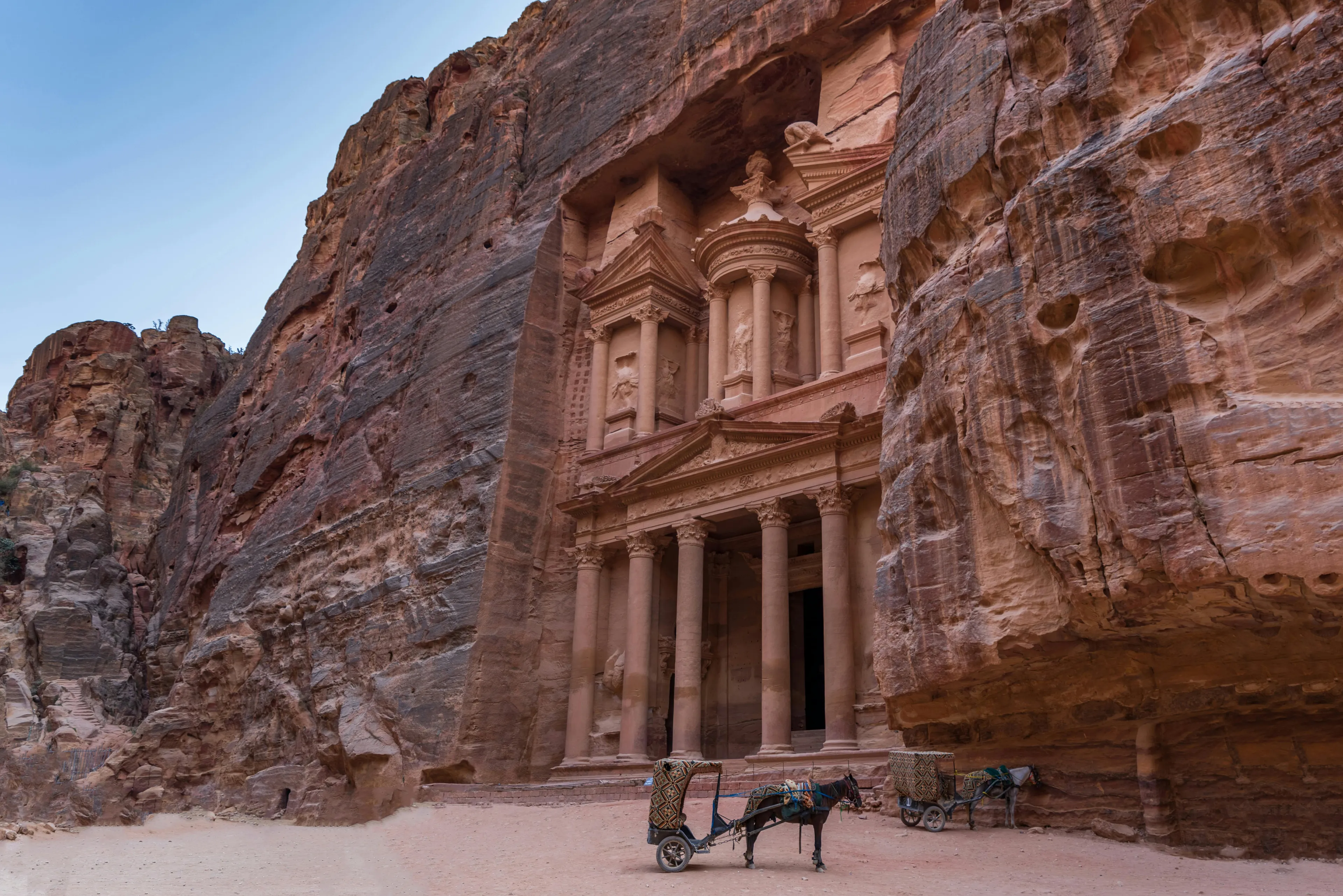 Ancient Nabataean city of Petra carved into red sandstone cliffs with horses and carts in the foreground.