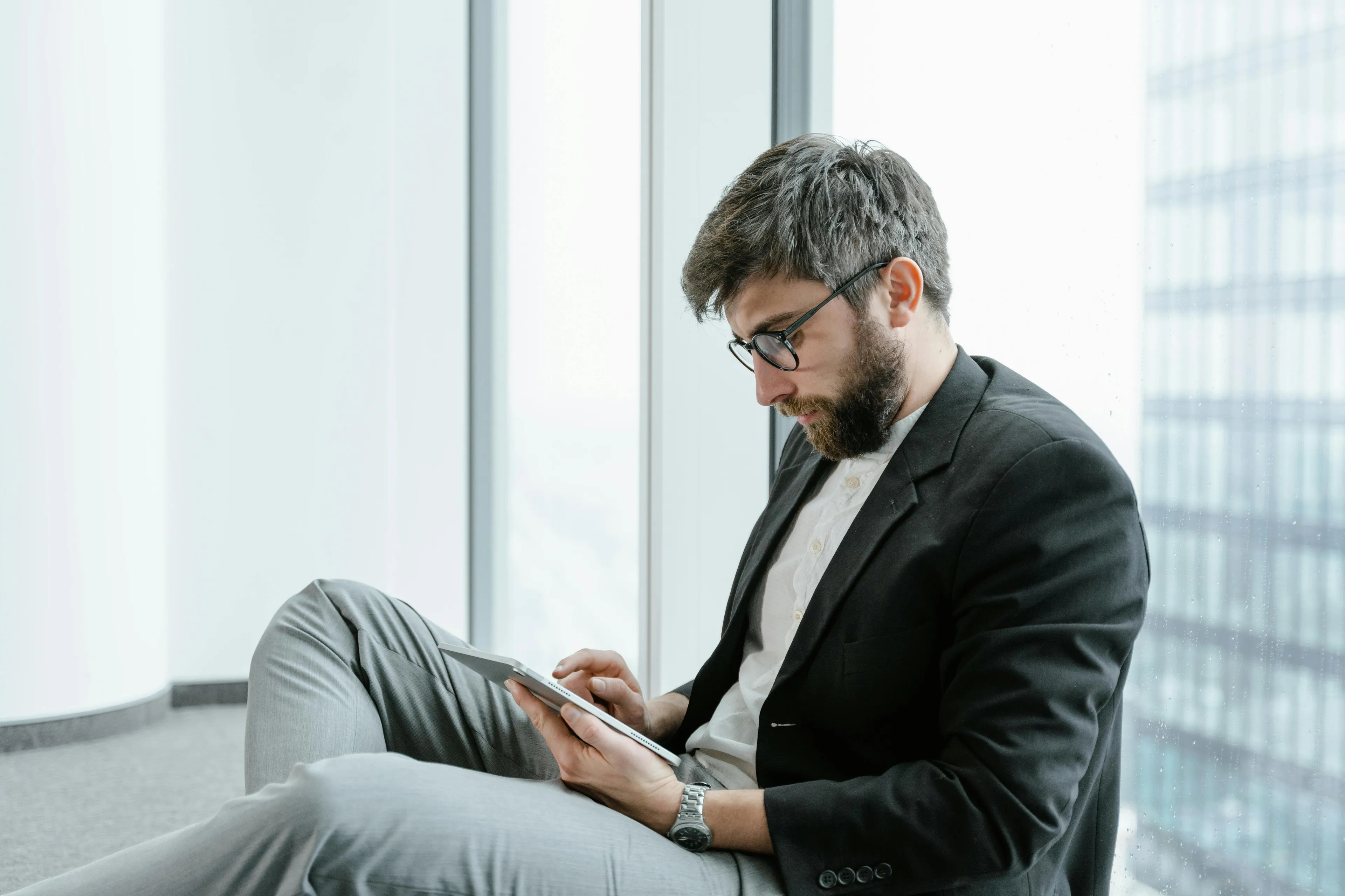 Man with glasses and beard in business attire sitting by a window, focused on a tablet for a vexillology quiz.