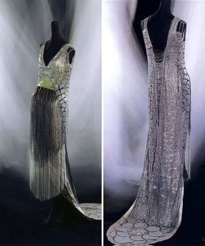 Silver and black historical garments with intricate beadwork and fringe details, displayed on mannequins against a white backdrop.