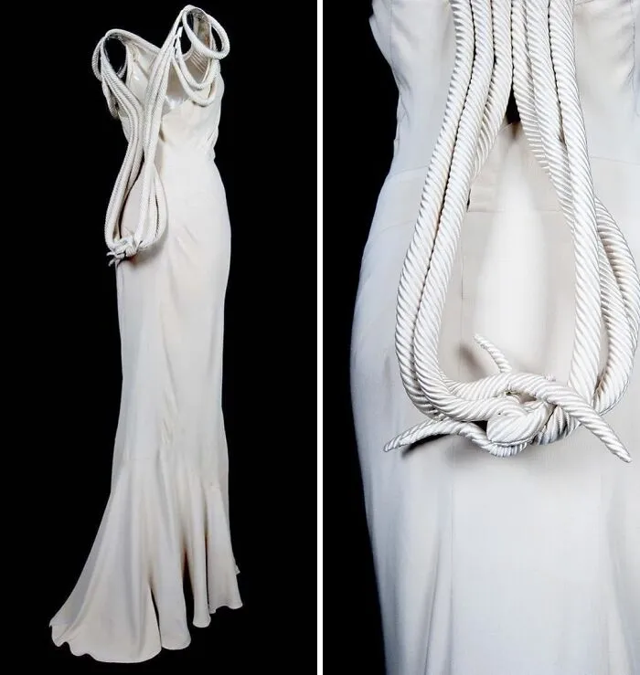 White historical garment with large rope detail draped over a long elegant dress on a black background.