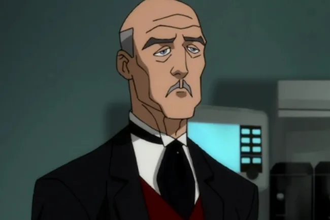 Alfred Pennyworth in a formal suit with a serious expression, standing in a dimly lit room with blurred background.