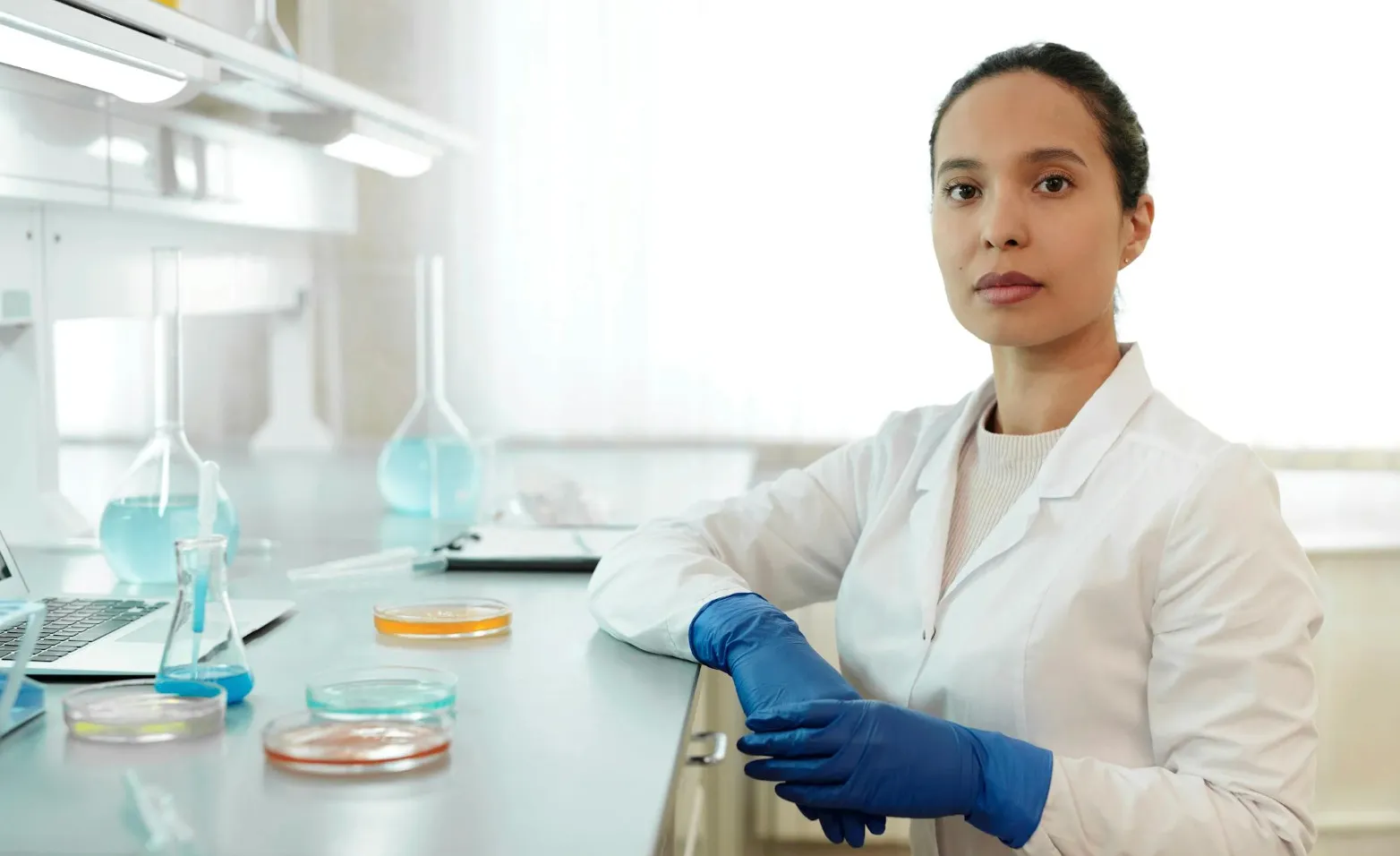 Scientist in a lab coat and blue gloves working at a counter with petri dishes and lab equipment for science quiz research.