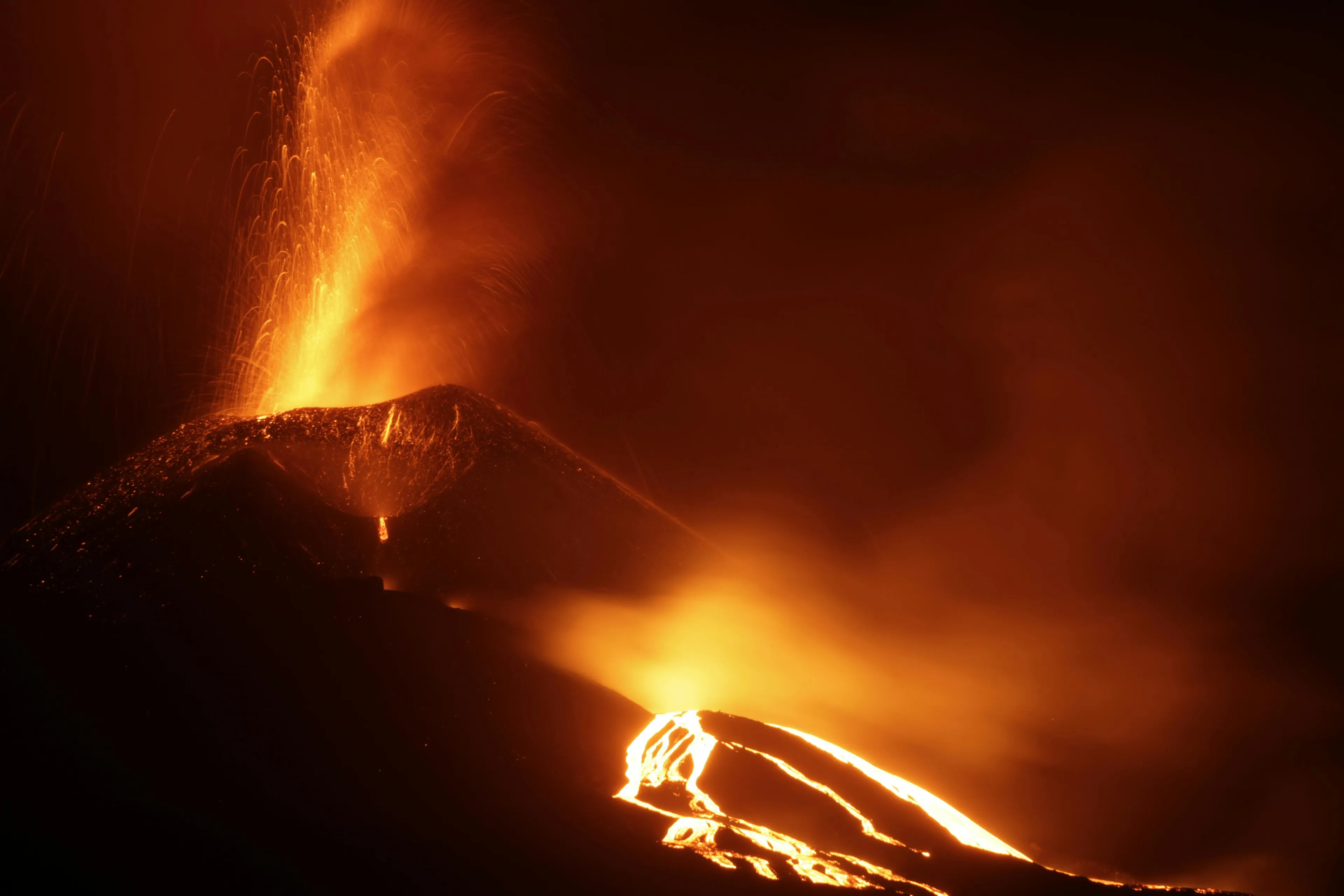 Volcanic eruption at night showing glowing lava flow and magma spewing from the crater in a fiery display.