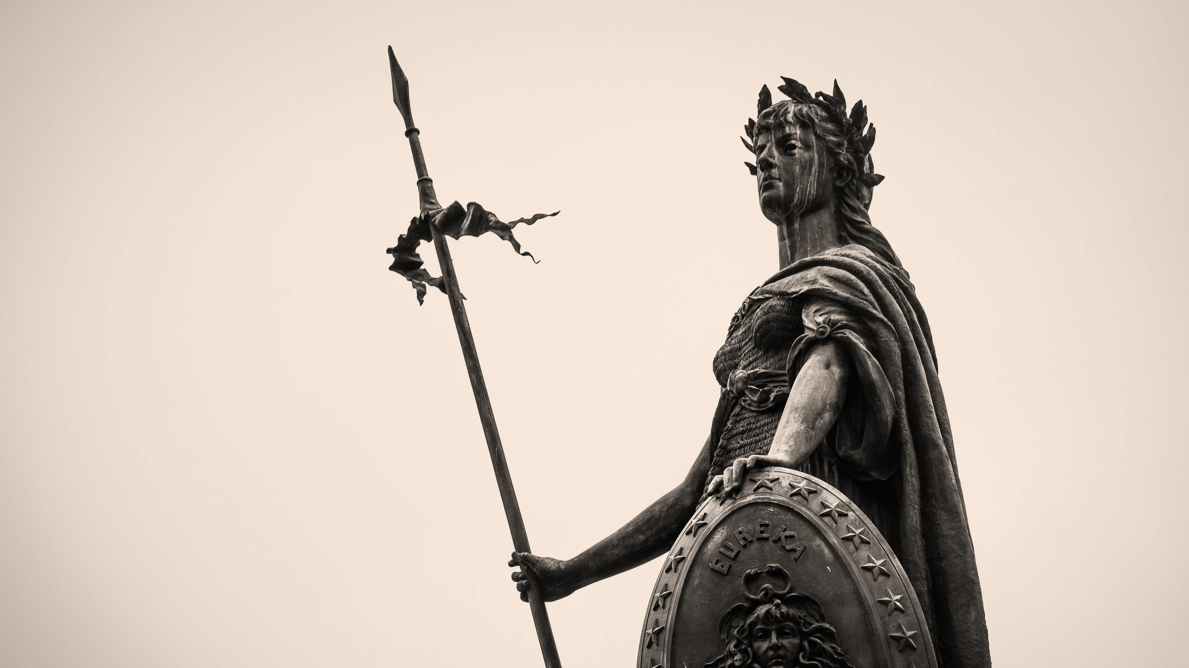 Black and white statue of a warrior holding a long pike with a decorated shield against a plain sky background.