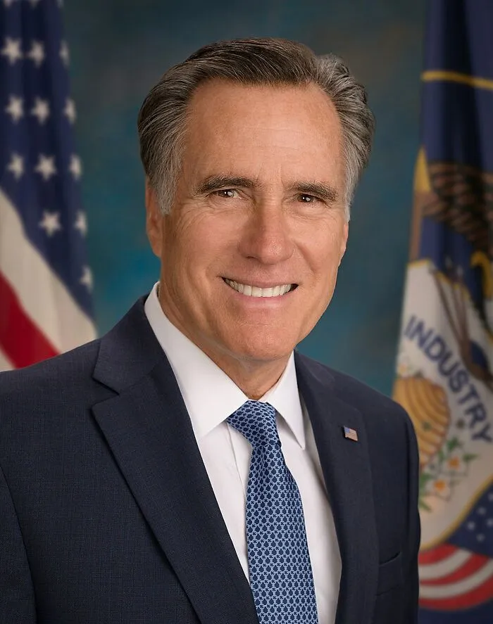 Middle-aged man in a suit and tie smiling in front of American flags, representing people who deserve an apology.
