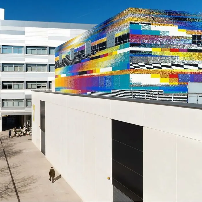 Edificio moderno con obra de arte urbano colorida y pixelada en la fachada superior bajo un cielo despejado azul.