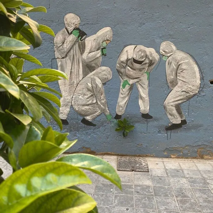 Arte urbano impresionante en pared, mostrando a figuras con trajes protectores cuidando una planta junto a hojas verdes.