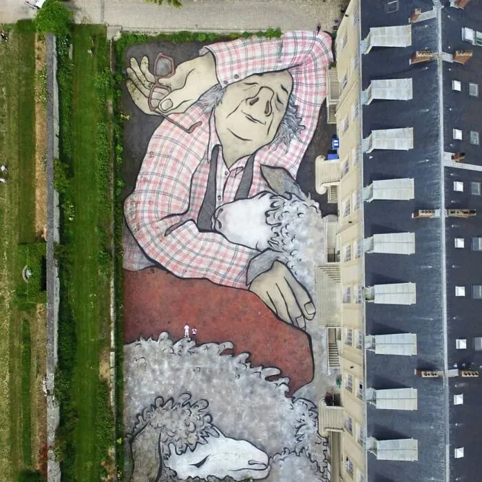 Obra de arte urbano gigante que muestra a un hombre abrazando ovejas, impresionantes detalles y colores vistos desde arriba.