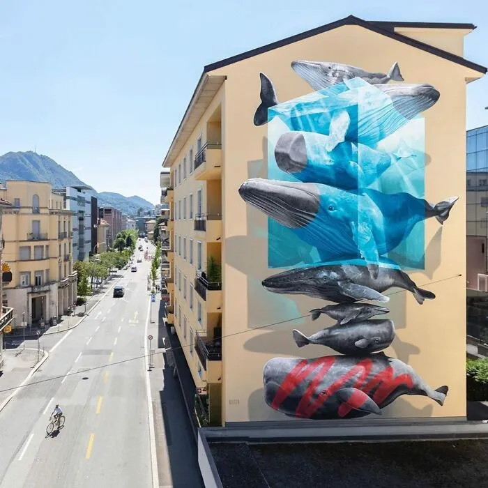 Mural de arte urbano en edificio mostrando ballenas azules en estilo tridimensional en zona urbana con montañas al fondo.