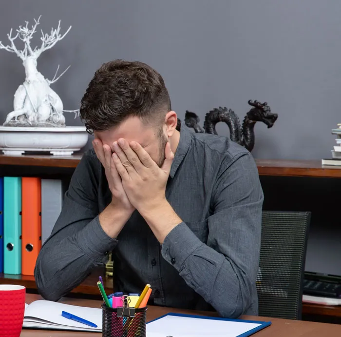 Man in gray shirt sitting at desk covering his face, illustrating emotional effects of dating out of their league experiences.