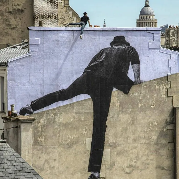 Hombre sentado en techo junto a impresionante obra de arte urbano en mural gigante que destaca en edificio antiguo.