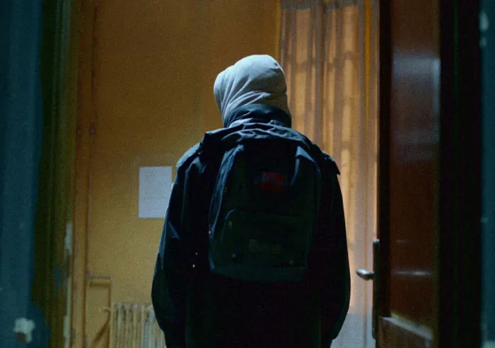 Person in a hoodie with a backpack standing in a dimly lit hallway, evoking mystery and intrigue themes.