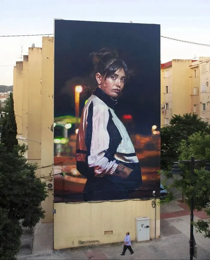 Mural de arte urbano con retrato realista de mujer joven en chaqueta deportiva en fachada de edificio alto.