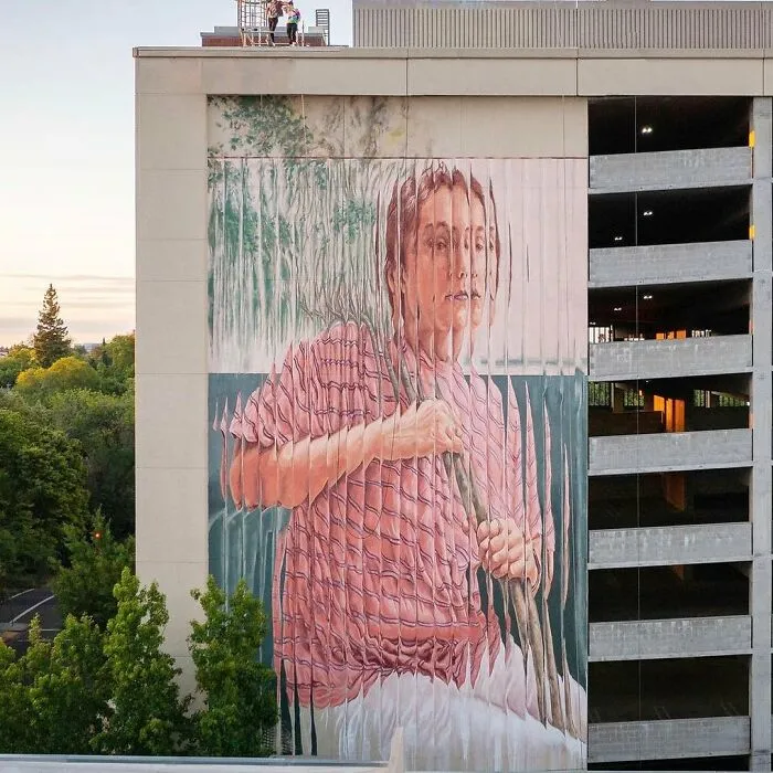 Mural de arte urbano impresionante en edificio alto con imagen de persona joven con camiseta rosa y fondo natural.