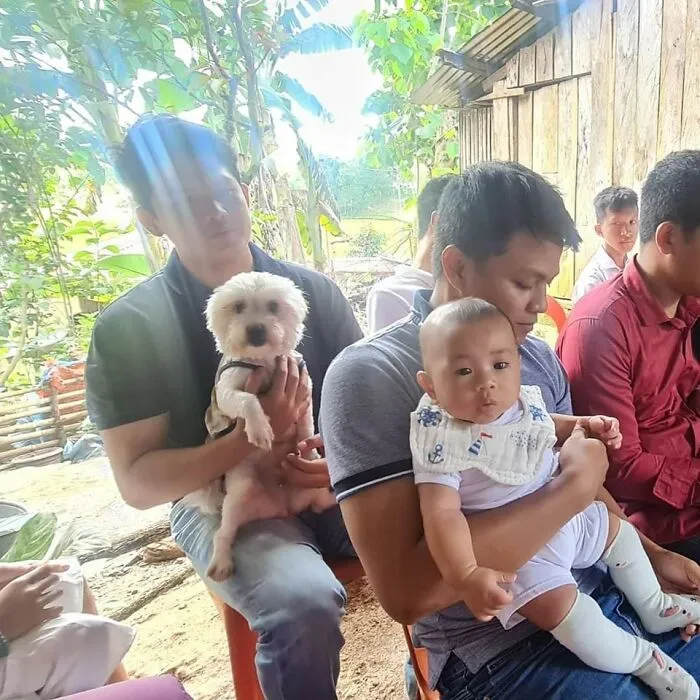 Two men sitting outdoors, one holding a small dog and the other holding a baby, showing unexpected beautiful bonds between humans and animals.