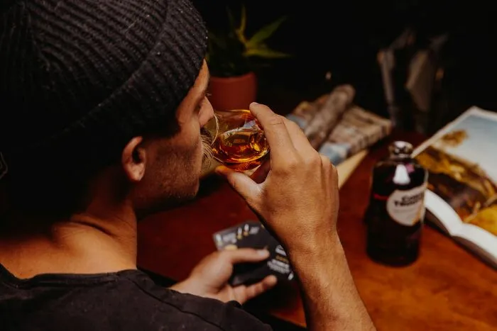 Man wearing a beanie drinking whiskey while holding cards, illustrating dangerous things that are often dismissed.