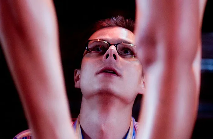 Young man wearing glasses looking upwards between two blurred arms, representing coworkers involved in workplace incidents.